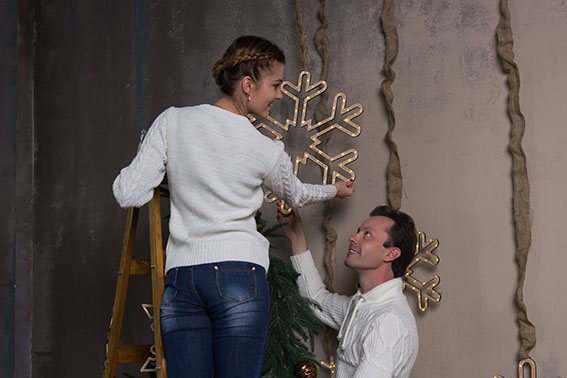 A young woman is standing on a stepladder and decorating a house for Christmas, and a man supports a ladder. A young couple decorates a house for Christmas with garlands of snowflakes