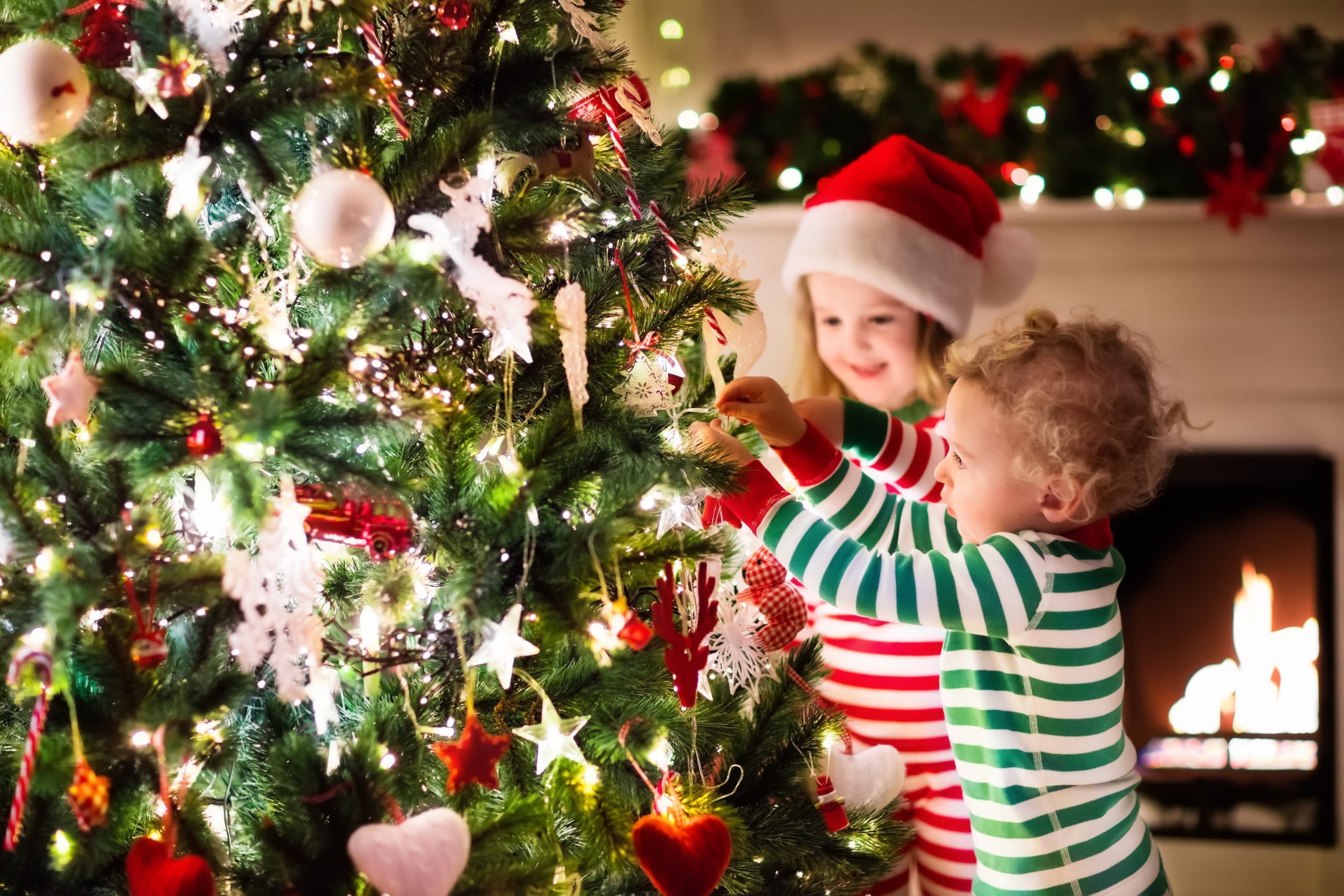 Happy little kids in matching red and green striped pajamas decorate Christmas tree in beautiful living room with traditional fire place. Children opening presents on Xmas eve.