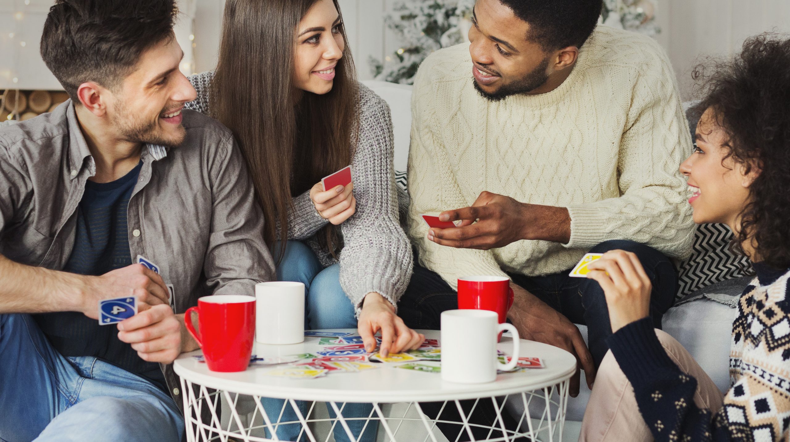 Multiracial friends having fun and playing game of cards UNO against christmas tree