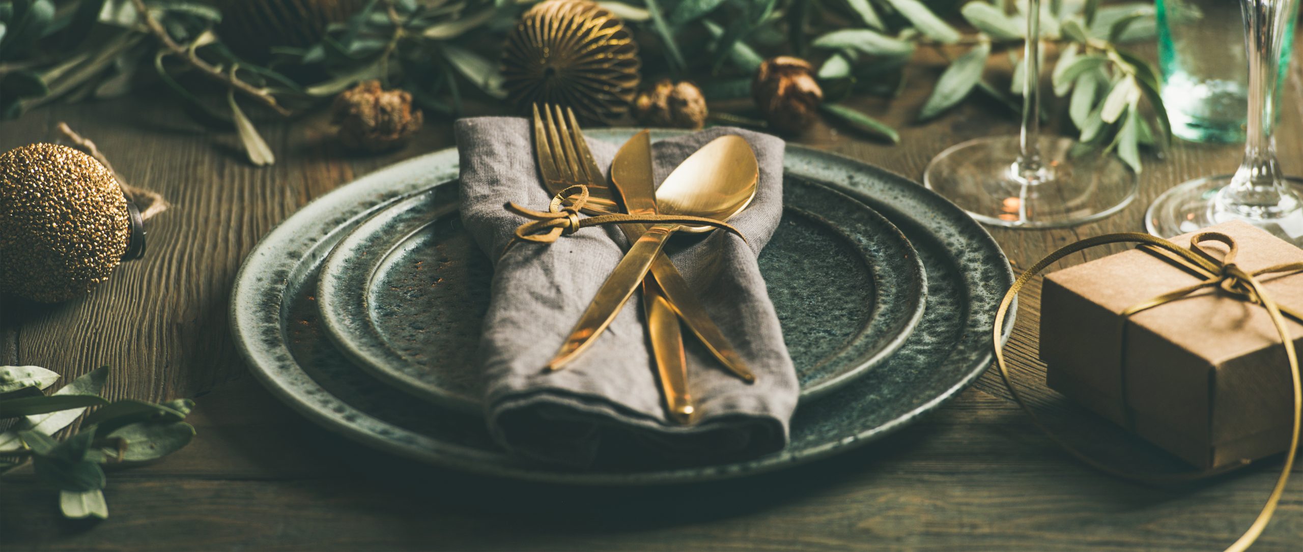 Christmas or New Years celebration party table setting. Plates, golden cutlery, glasses, gift box, festive branch decoration, candles, gliterring toys over rustic wooden table background