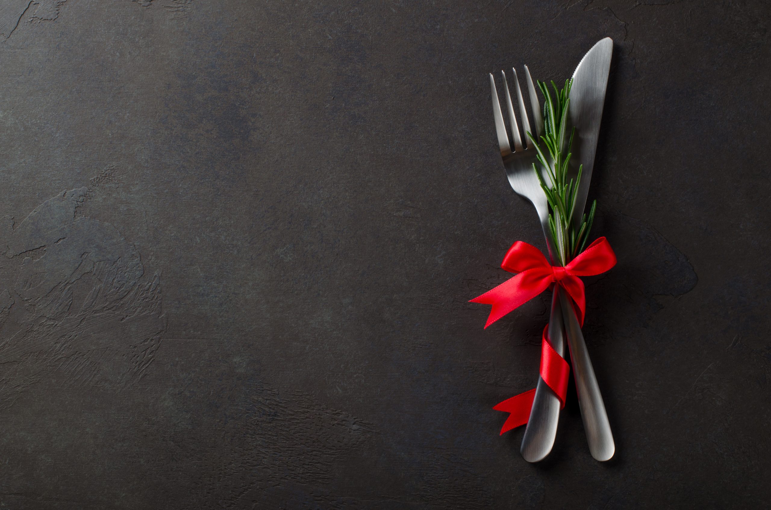 Festive set of knife and fork with red satin bow with rosemary, dark stone slate background, top view, copyspace, horizontal image