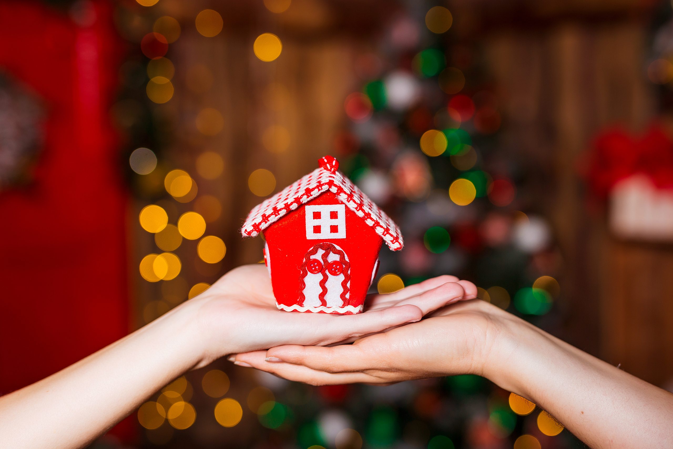Human hands holding decorative red house against blurred background. Christmas and home comfort concept
