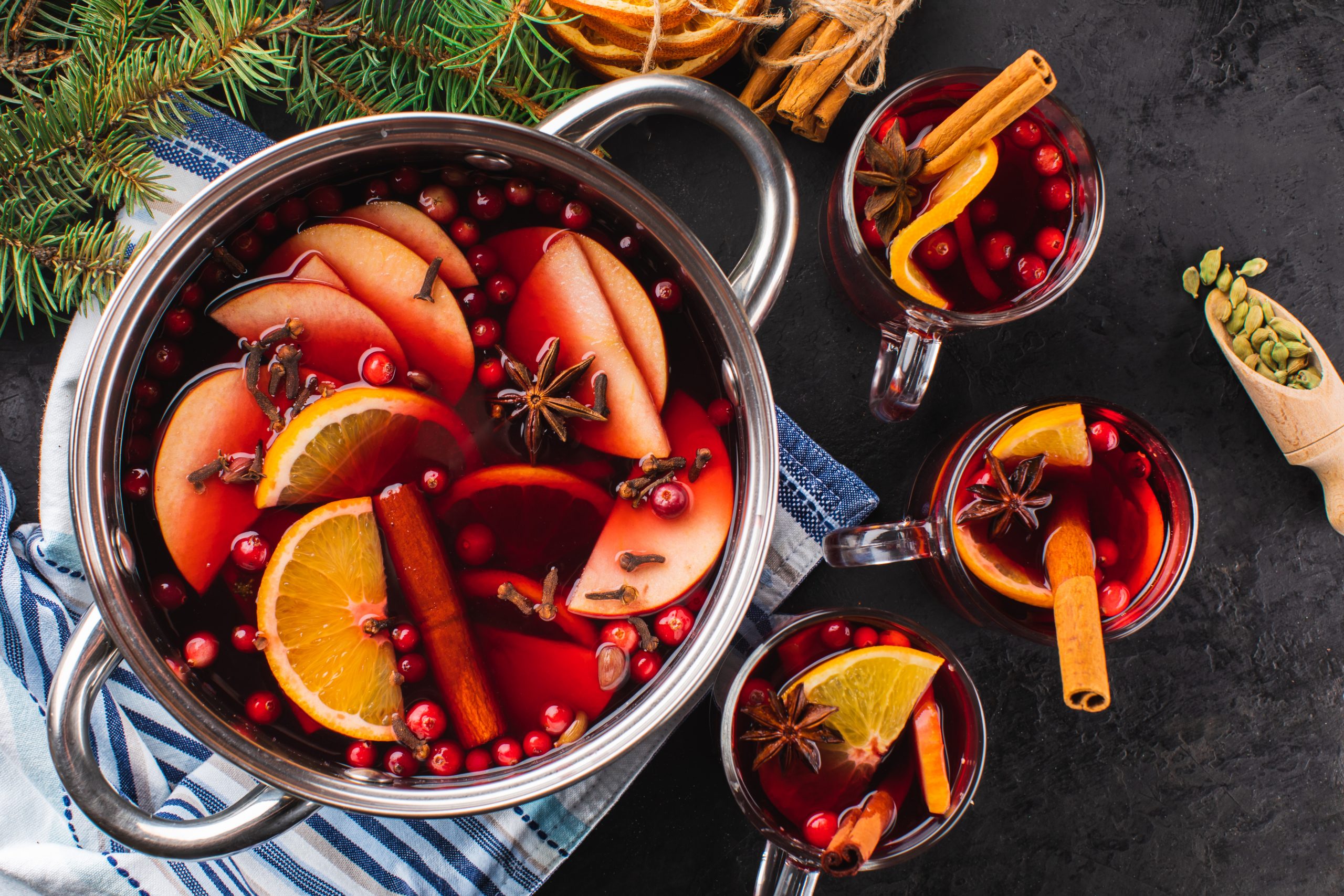 Mulled wine or punch in a glass and hot wine in a cooking pot with spices and fruits on a black textured background, top view. Christmas hot drink.